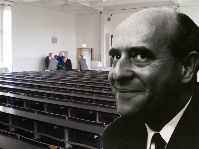 Im Bild zu sehen ist ein Hörsaal. Am Rand der ersten Reihe stehen fünf Personen zusammen. Rechts im Bild ein Porträt von Hans Blumenberg.